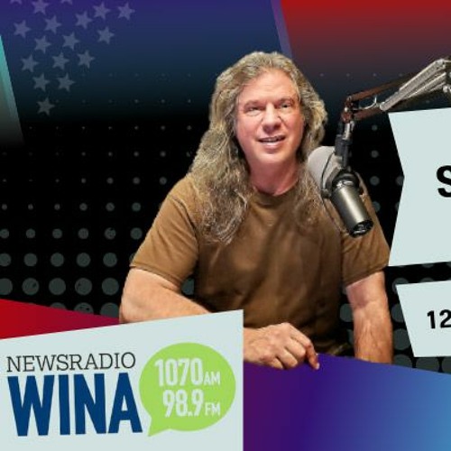Man with long hair sitting at a radio studio desk with a microphone, smiling.
